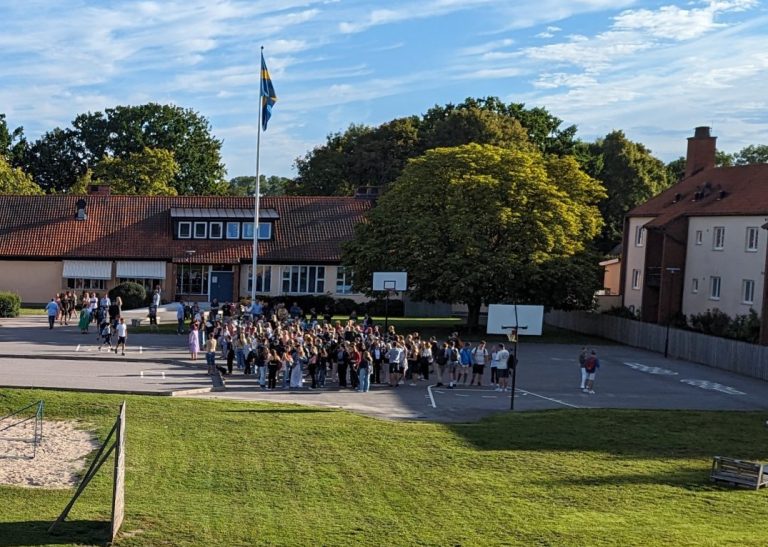 Foto av en skolgård med grönområden, sandlåda och bollplan. En grupp elever står samlad framför skolbyggnaderna, som har solpaneler på taken. En svensk flagga vajar på flaggstången under en klarblå himmel.
