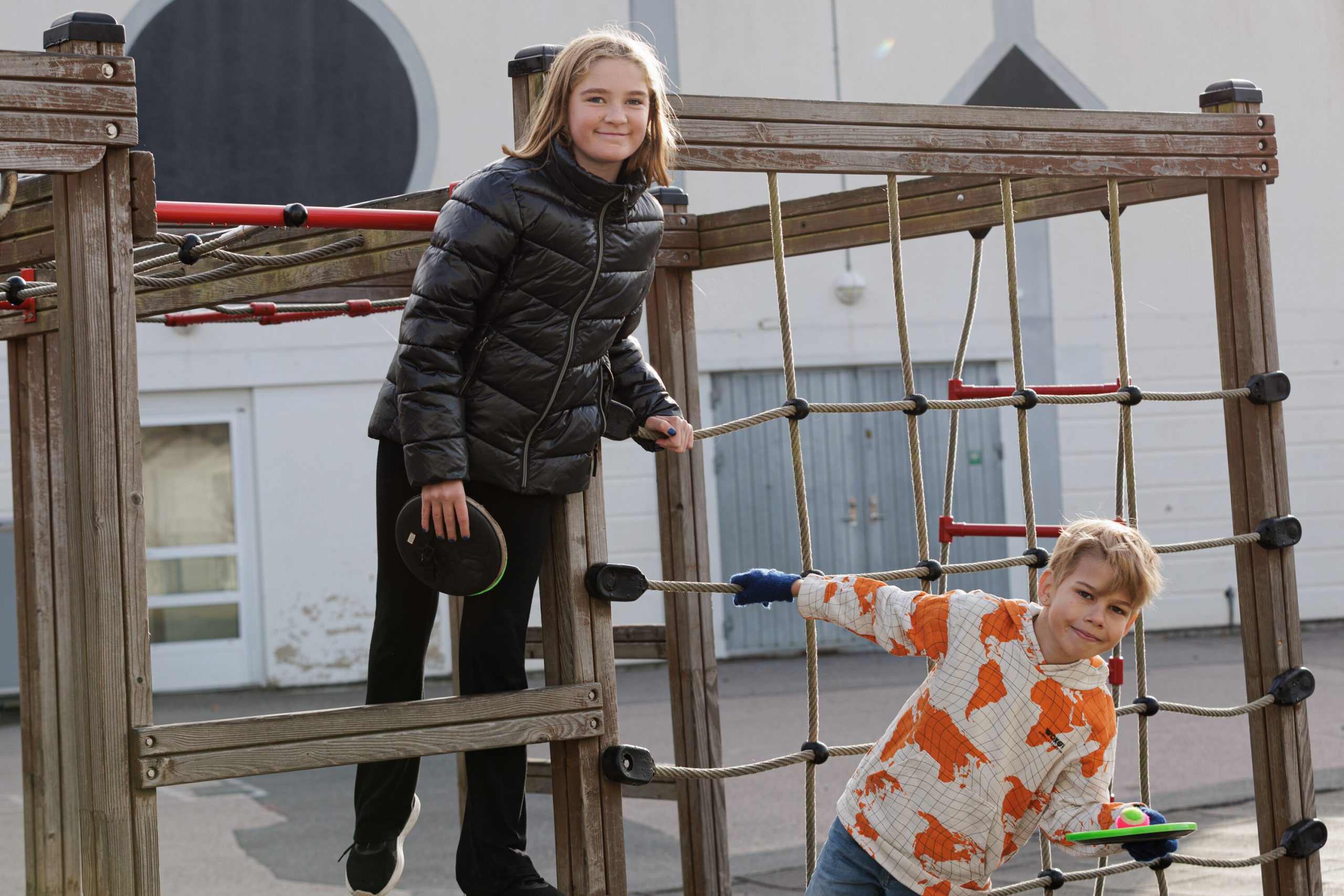 Två barn leker på en klätterställning av trä och rep på en skolgård en solig dag. Flickan i svart jacka står högre upp och håller i en liten racket, medan pojken i mönstrad tröja klänger sig fram i repen längre ner, båda ler mot kameran.