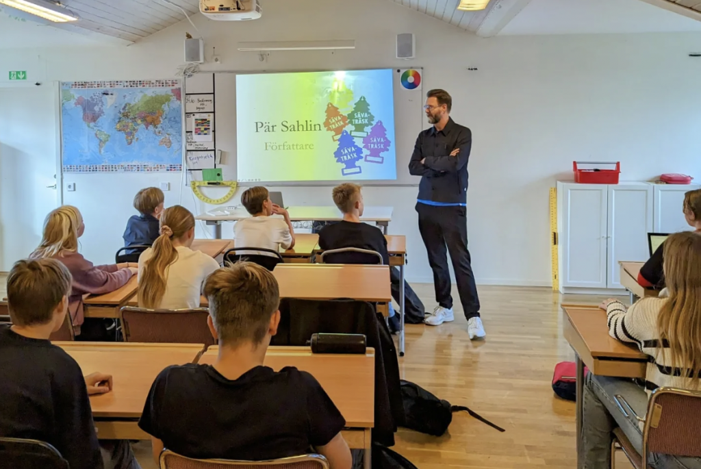 En författare håller en presentation för en klass, med en presentationsslide som visas på en skärm i bakgrunden. Eleverna sitter vid sina bänkar och lyssnar i ett ljust klassrum med en världskarta på väggen.